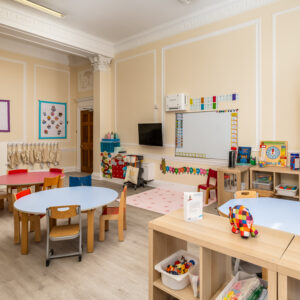 EIFA In a brightly decorated classroom at EIFA in London, round tables and small chairs invite young learners. A whiteboard, colorful bilingual decorations, and shelves with toys and books fill the room. Educational posters adorn the walls, while a TV is mounted above the board.