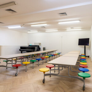 EIFA A brightly lit room with long folding tables and colorful round stools in EIFA London. A black piano graces the back wall, while a large flat-screen TV is positioned off to the side. The light wood-patterned floor and fluorescent-lit ceiling create a welcoming bilingual learning space.