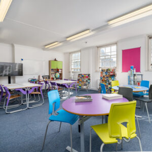 EIFA The EIFA classroom features a blue carpet, circular tables, and colorful chairs. A bilingual learning environment is enhanced with a large whiteboard and TV. Shelves by the window hold books and supplies, while bright lighting and a pink bulletin board add to the lively atmosphere.