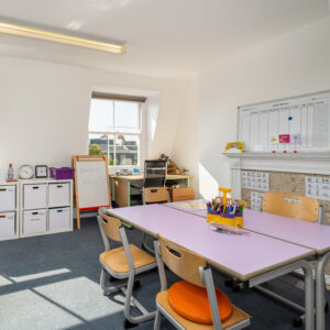 EIFA A bright classroom at the EIFA School features a purple table, wooden chairs, and various educational materials. The bilingual schedule is displayed on a whiteboard, while shelves with storage boxes line the walls. A window lets in natural light, beautifully illuminating the space.