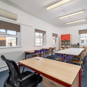 EIFA A bright classroom in a London school, with several desks and chairs arranged in rows. A teachers desk and chair are in the foreground. The room has large windows on the left, a bookshelf with colorful storage, and fluorescent ceiling lights.