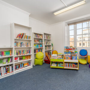 EIFA A brightly lit library corner with white bookshelves filled with books sits in a bustling London school. Colorful bean bags and low yellow book racks showcase more books near a large window, offering views of city buildings. The blue carpeted floor adds a cozy touch to this inviting bilingual space.