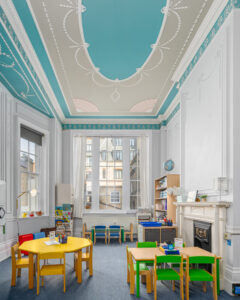 EIFA A bright London classroom at EIFA boasts ornate blue and white ceilings and large windows. Yellow and green tables, surrounded by small chairs, fill the space alongside bookshelves and a fireplace. The walls are adorned with intricate moldings and decorations, creating a charming school atmosphere.