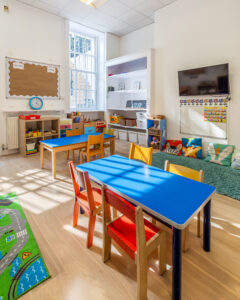 EIFA A brightly lit bilingual classroom in London features two blue tables, small colorful chairs, a play mat, a TV on the wall, and shelves filled with toys. The cozy reading area with cushions is bathed in sunlight streaming through a large window, creating a welcoming atmosphere.