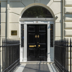 EIFA Elegant black double-door entrance with gold fixtures and the number 36 above, welcoming visitors in a bilingual setting. Flanked by narrow black railings and topped with an arched window. White columns and decorative masonry frame the doorway of EIFA School, set against a beige building exterior.