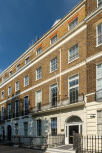 EIFA A four-story building in London with a classic brick facade features large white-framed windows. The ground floor is painted cream, and the entrance is arched. A black iron fence lines the front, and the sky is clear and blue, complementing this bilingual citys charm.