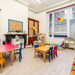 EIFA A bright, tidy preschool classroom in London with colorful tables and chairs. Shelves hold toys and learning materials. Large windows let in natural light. Decorations include balloons and a patterned ceiling border, reflecting the bilingual environment of EIFA. A cozy fireplace is also present.