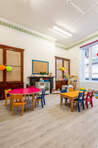 EIFA A bright and colorful bilingual classroom at EIFA with small tables and chairs in red, yellow, and blue. The room features playful decorations, a fireplace, and large windows for natural light. Educational toys and materials are neatly arranged in this inviting school environment.