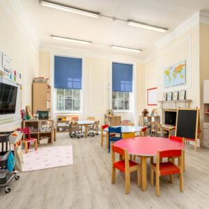 EIFA A bright and spacious classroom at EIFA in London, featuring wooden flooring, colorful round tables with small chairs, a chalkboard, and shelves with books and toys. A map adorns the wall alongside large windows with blue curtains, fostering a bilingual learning environment.