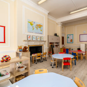 EIFA A bright, tidy bilingual classroom at EIFA with light-colored walls features a world map above a fireplace, round tables with colorful chairs, bookshelves filled with educational materials, and childrens artwork. The well-organized school room is vibrant and inspiring.