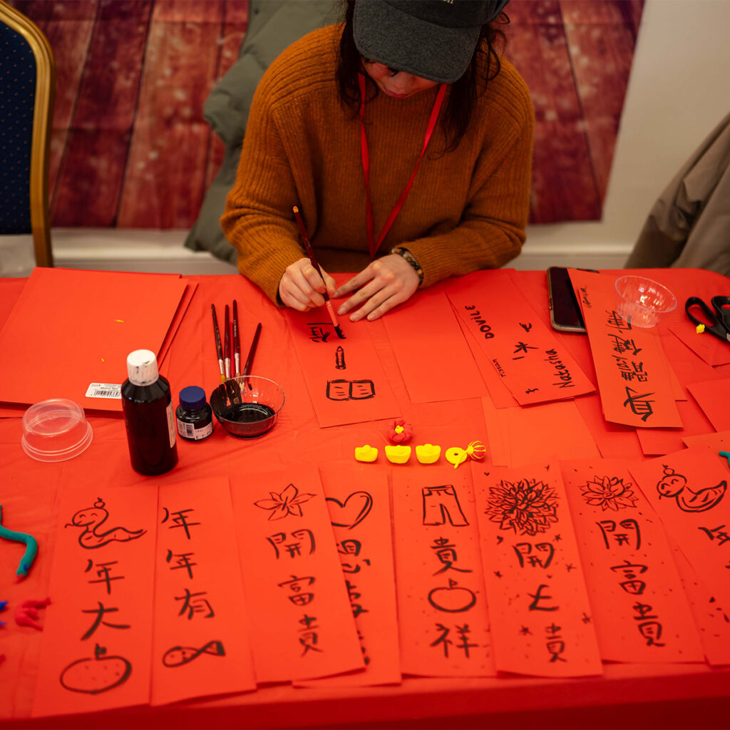 EIFA A person sits at a red table, practicing Chinese calligraphy with ink and brush. The table is covered with red paper sheets featuring various Chinese characters and symbols. Small decorative items are also on the table.