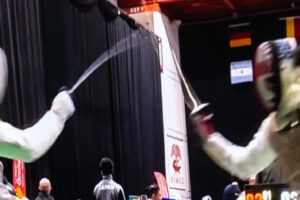 EIFA Two fencers in protective gear and masks compete on a platform, surrounded by flags from various countries. The indoor scene, akin to an intense treasure hunt at the National Gallery, unfolds against a dark curtain, emphasizing the duel between the athletes.