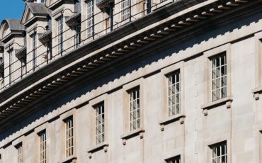 EIFA Façade incurvée dun bâtiment historique avec des fenêtres régulièrement espacées sur un ciel bleu clair. Larchitecture présente des détails en pierre et une rangée de lucarnes le long de la ligne de toit. Un autre bâtiment est partiellement visible sur la gauche.