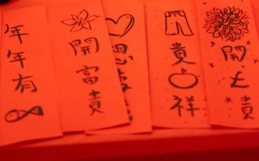 EIFA Red paper strips with Chinese calligraphy and drawings, featuring symbols like a fish, apple, heart, and flower. Bright clay pieces spelling SNAK rest on the table amid celebratory decorations for a festive occasion. Its like a treasure hunt of vibrant art at the National Gallery!.