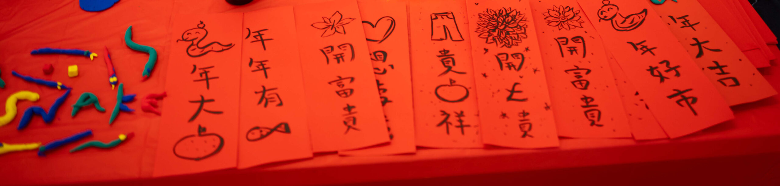 EIFA Red paper strips with Chinese calligraphy and drawings, featuring symbols like a fish, apple, heart, and flower. Bright clay pieces spelling SNAK rest on the table amid celebratory decorations for a festive occasion. Its like a treasure hunt of vibrant art at the National Gallery!.