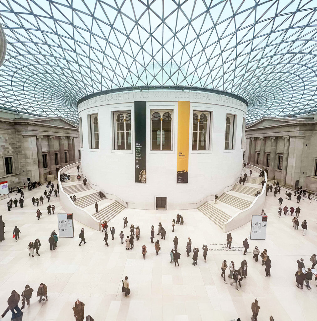 EIFA People walk through a spacious atrium with a large circular white structure at the center, under a geometric glass roof. Stairs lead up to the structure, which has banners hanging on its side. The architectural space is bright and airy.