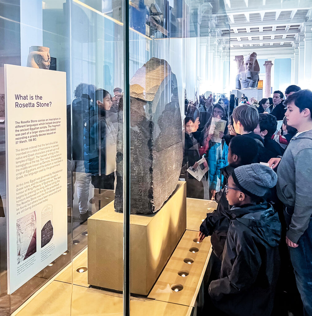 EIFA A diverse group of people, including children and adults, gather around the display case of the Rosetta Stone in a museum. The Stone is upright, surrounded by informational displays. The room has large windows and bright lighting.