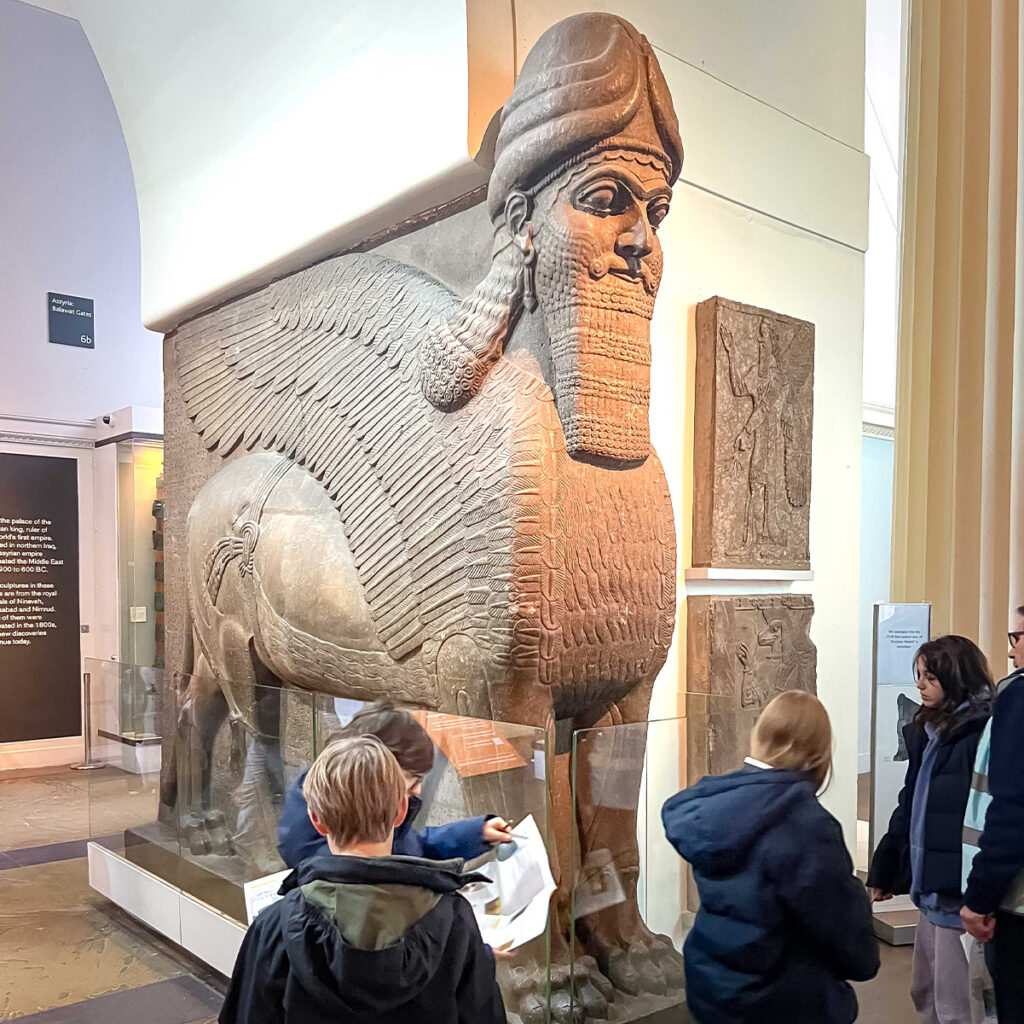 EIFA A large stone statue of a winged, bearded figure with a human head and lions body stands in a museum. Several visitors, including children, are observing it closely. The surrounding walls feature additional carvings and exhibits.