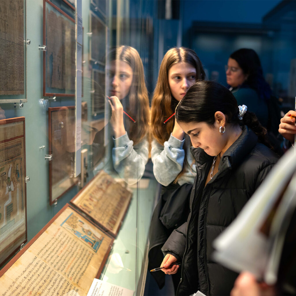 EIFA People are closely examining historical documents displayed in a glass case at a museum. Two young women are in the foreground, one holding a notepad. There are reflections on the glass, and more visitors are blurred in the background.