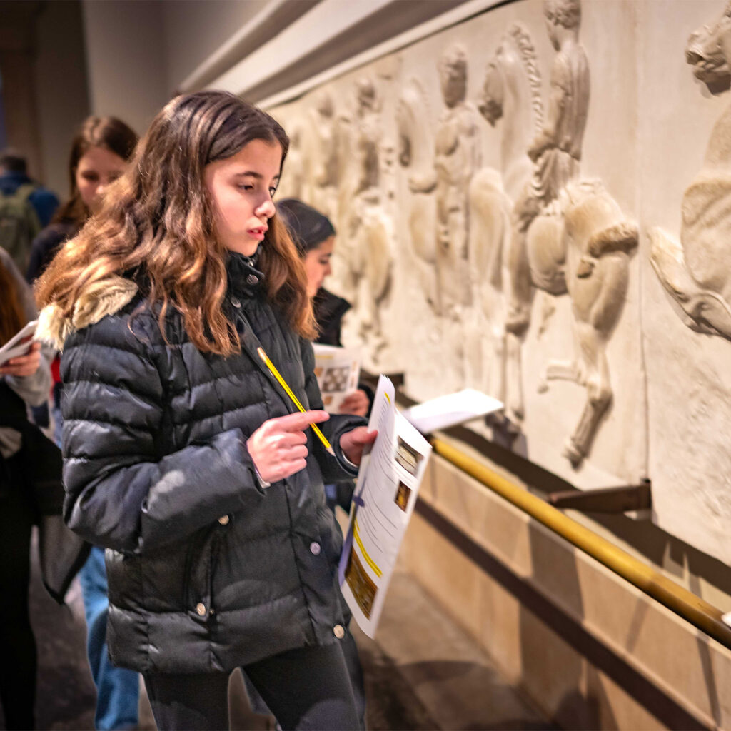 EIFA A young girl in a black jacket observes a historical frieze inside a museum. She holds a booklet and a pencil, suggesting shes taking notes. Other visitors are in the background, also engaged with the exhibits.