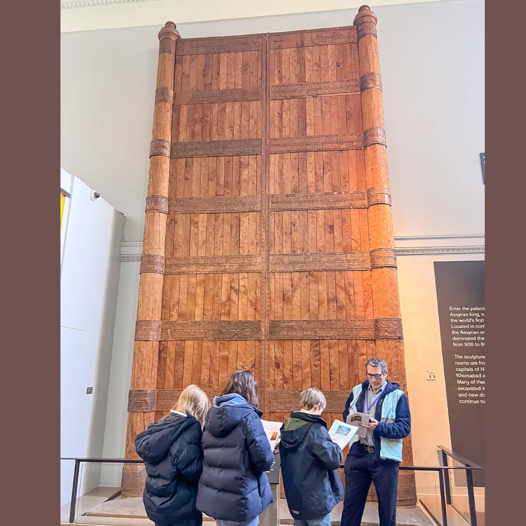 EIFA A group of people stand in front of a large, ancient, wooden door or gate displayed in a museum. The door features intricate carvings and towering vertical panels. Plaques with information are visible nearby.