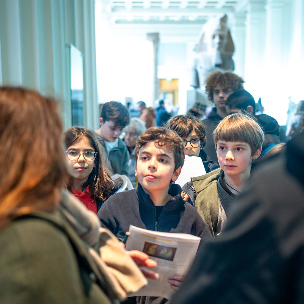 EIFA A group of children holding papers are attentively looking at a guide inside a museum. Statues and architectural details are visible in the background, creating a cultural setting.