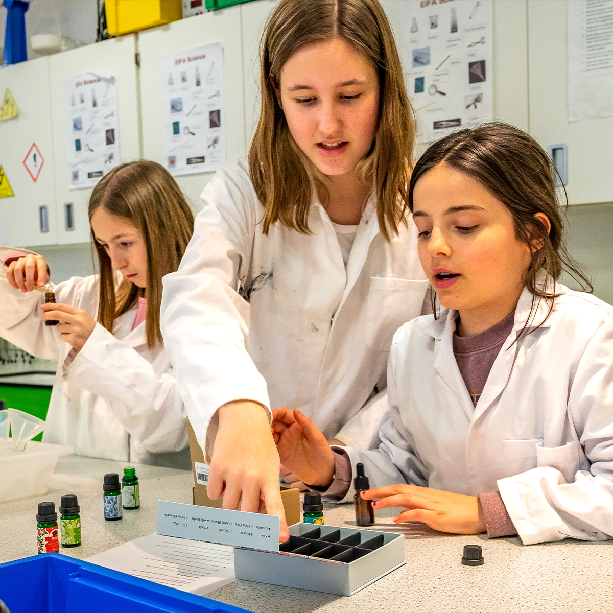 EIFA Three bilingual children in white lab coats conduct a science experiment. One guides the others using a box with compartments, while another examines a vial. In the background, the EIFA London science lab setup features educational posters and advanced equipment.