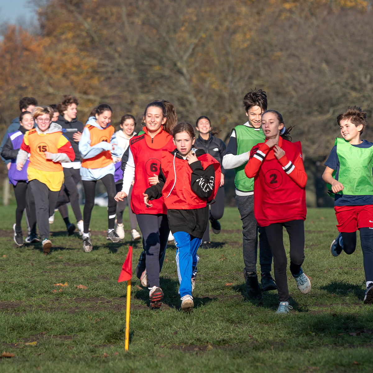 EIFA A group of older children from EIFA School is running in a grassy park during a sports event. Wearing colorful, numbered sports bibs—with red and green prominent—they dash past trees with autumn leaves under the clear sky, showcasing their bilingual spirit through energetic teamwork.