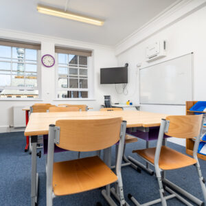 EIFA An empty classroom with wooden desks and chairs, a whiteboard, and a wall-mounted TV. The clock reads 10:10 on the white wall. Large windows offer views of a building and tower in London. The blue carpet contrasts with colorful bins in the corner, reflecting the vibrant EIFA bilingual learning environment.