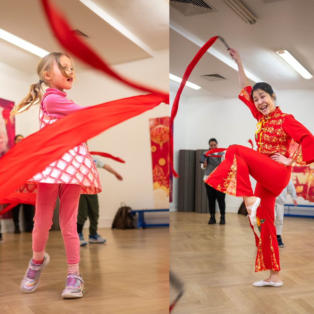 EIFA A girl and a woman dance energetically with red ribbons in a bright room. The girl wears pink, and the woman wears a red outfit with gold details, both smiling and in mid-movement. There are onlookers in the background.