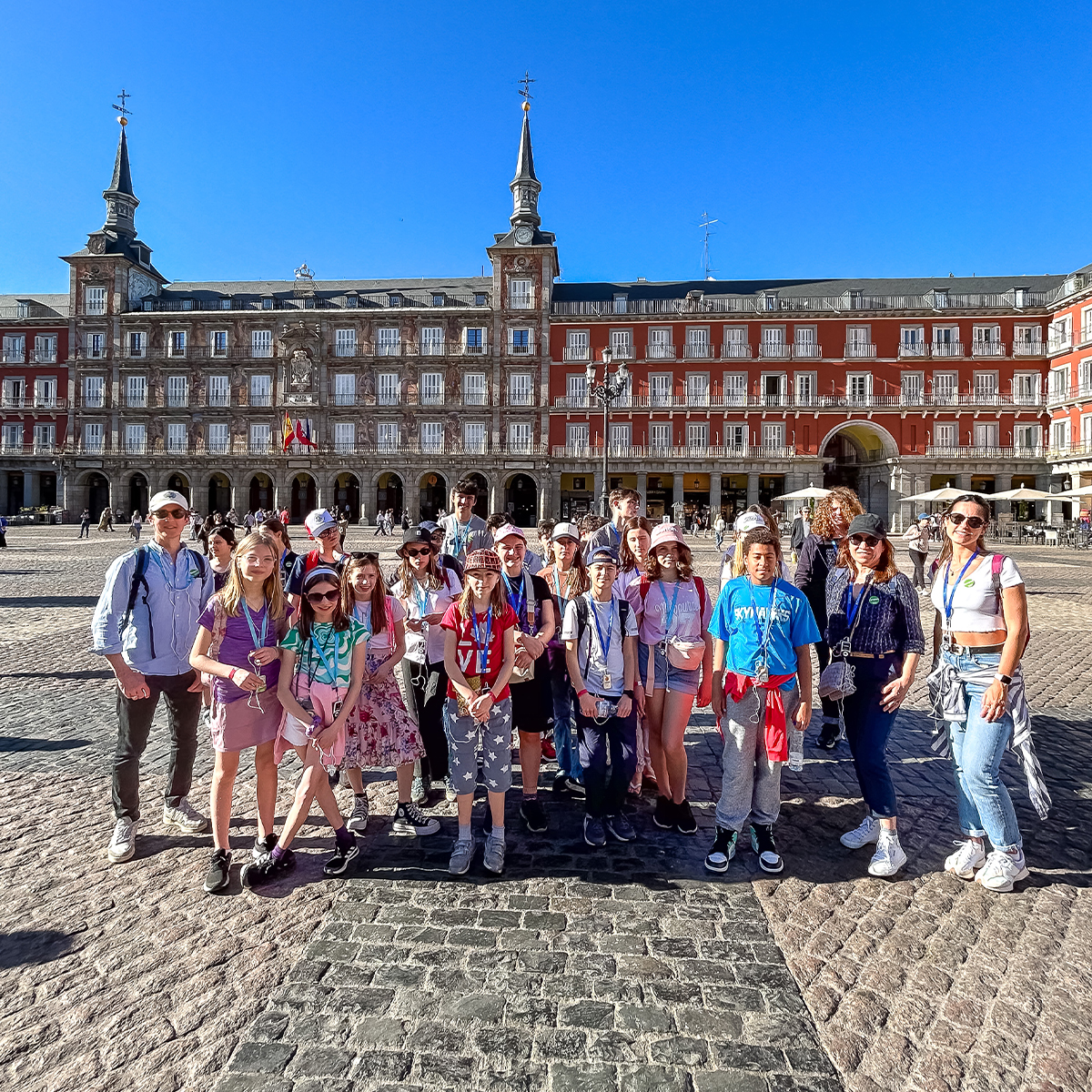 EIFA A diverse group of people, including children and adults, stand together smiling in an open plaza with a historic building in the background. Beneath the clear blue sky, the bilingual gathering takes place on cobblestones, embodying the vibrant spirit of London.