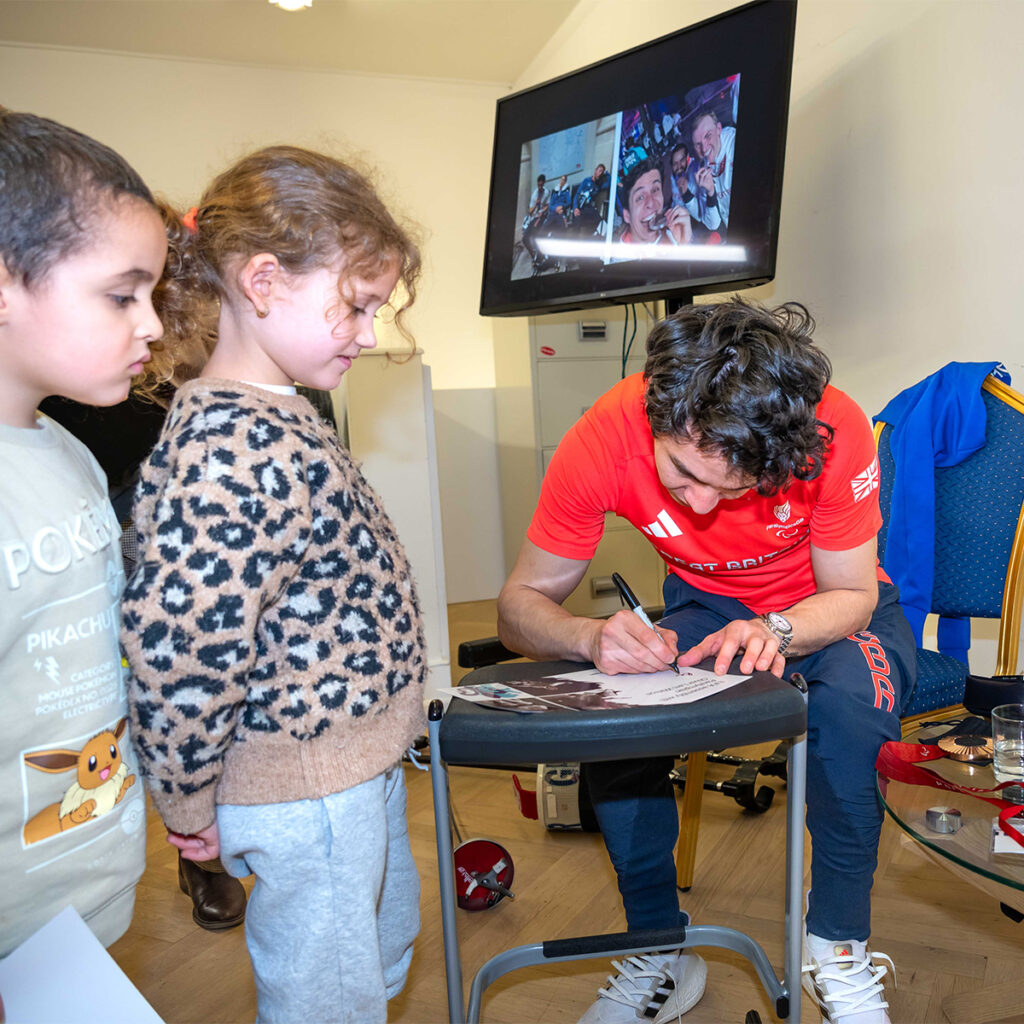 EIFA A person in a red shirt signs an autograph for two young children. A TV screen in the background displays images of smiling people. The room appears casual, with chairs and a table nearby. The children watch attentively.