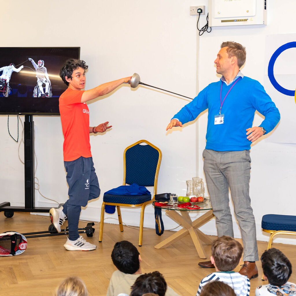 EIFA A person in a red shirt playfully holds a fencing sword near a man in a blue sweater, who raises his arms in mock surrender. They stand in a room with children seated on the floor watching. A TV nearby shows a fencing scene.