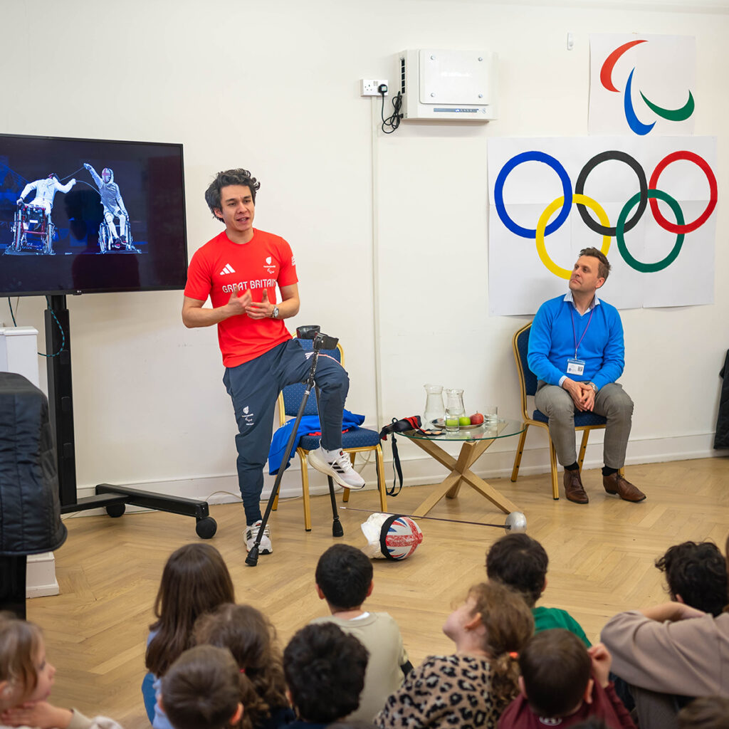 EIFA A person with a prosthetic leg, wearing a red shirt, stands speaking to a group of children in a classroom. A screen displays a sports image. A seated person listens nearby. Olympic and Paralympic logos are on the wall.