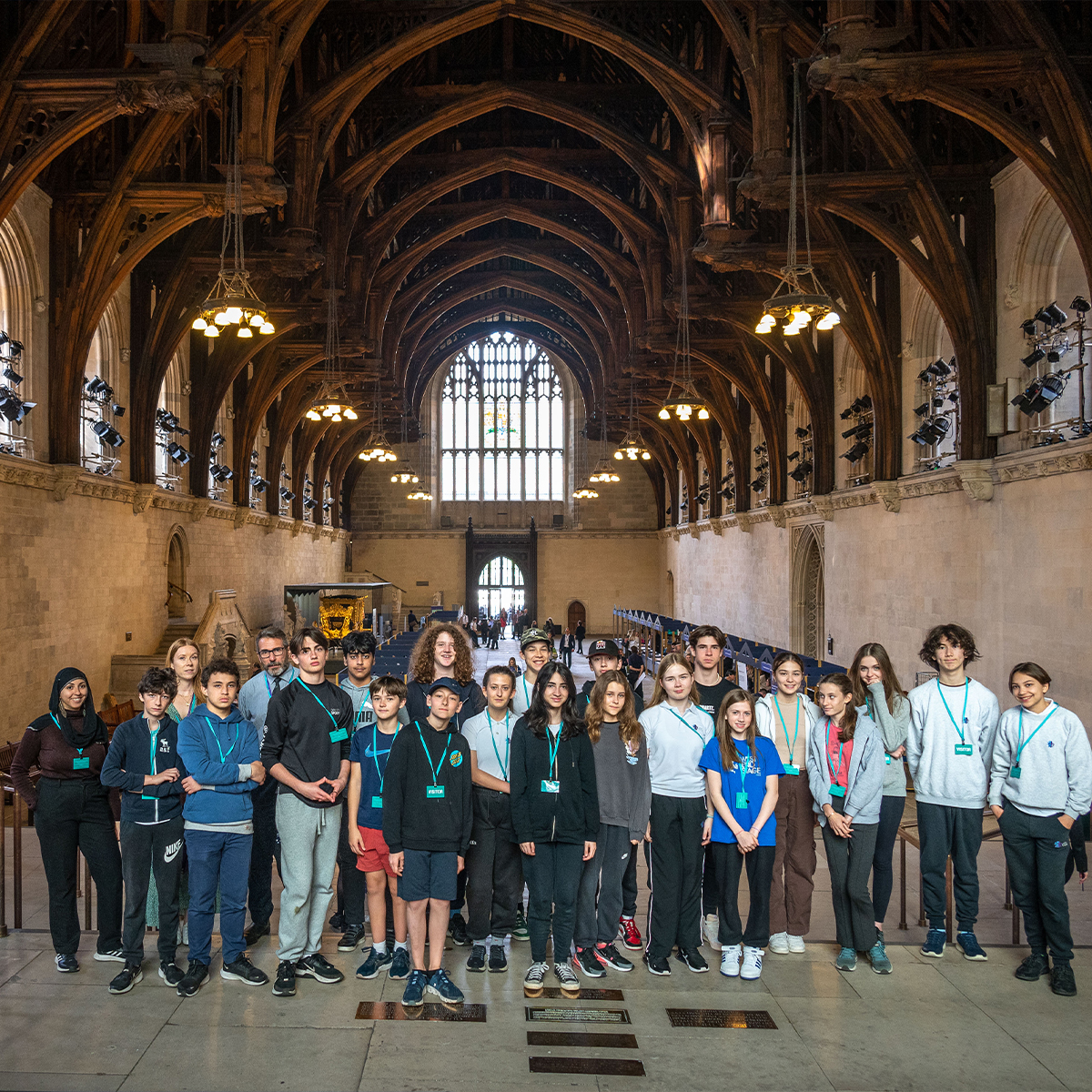 EIFA A group of young people stands together inside a large hall with a high, arched wooden ceiling and a grand stained glass window in the background. Some wear blue lanyards bearing the EIFA logo as they pose in a single row. The spacious, well-lit hall reflects Londons blend of history and modernity.