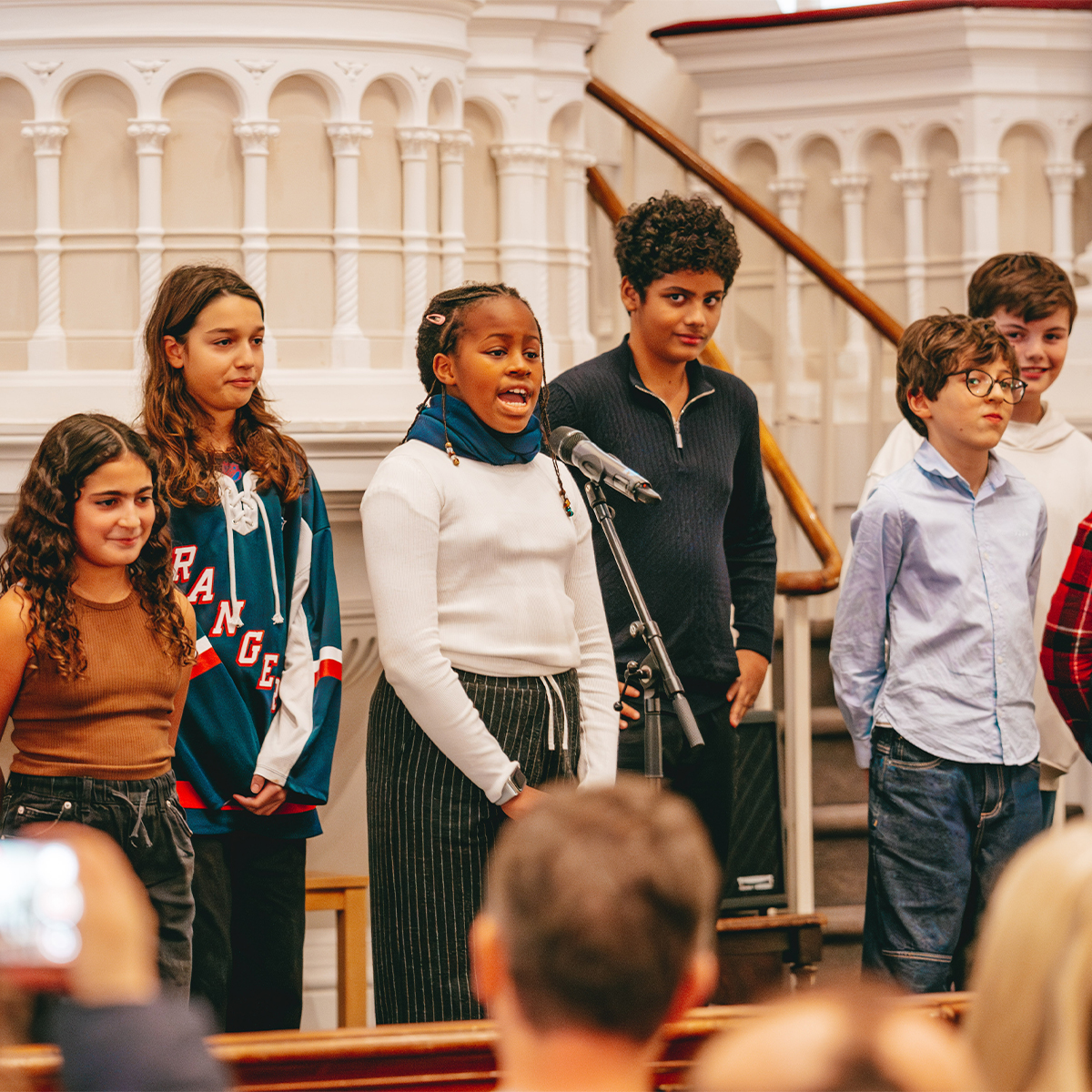 EIFA A group of six bilingual children stands on a stage in London, gathered around a microphone. One child is speaking confidently. The setting features ornate white architectural elements, with an engaged audience partially visible in the foreground, capturing the essence of EIFAs spirit.