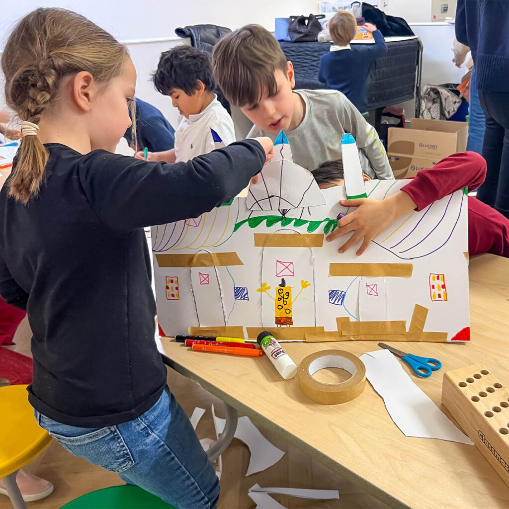 EIFA Children are crafting a cardboard house, using paper, markers, glue, and tape. They are gathered around a table focused on assembling and decorating the structure. Various art supplies are scattered around them.