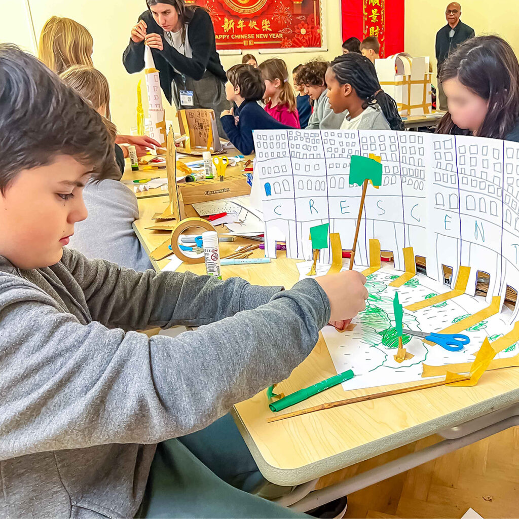 EIFA Students sitting at tables work on constructing paper models of buildings labeled CRESCENT. Supplies like scissors and glue are on the table. A teacher assists in the background, and Chinese New Year decorations are visible.