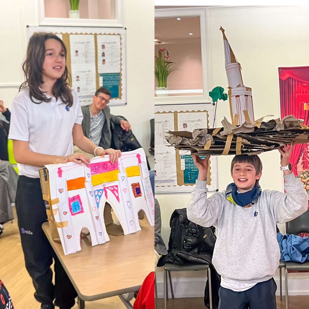 EIFA Two children presenting creative projects in a classroom. The girl on the left holds a colorful paper castle made from cardboard. The boy on the right raises a large, circular paper structure resembling a tower with a green decoration on top.