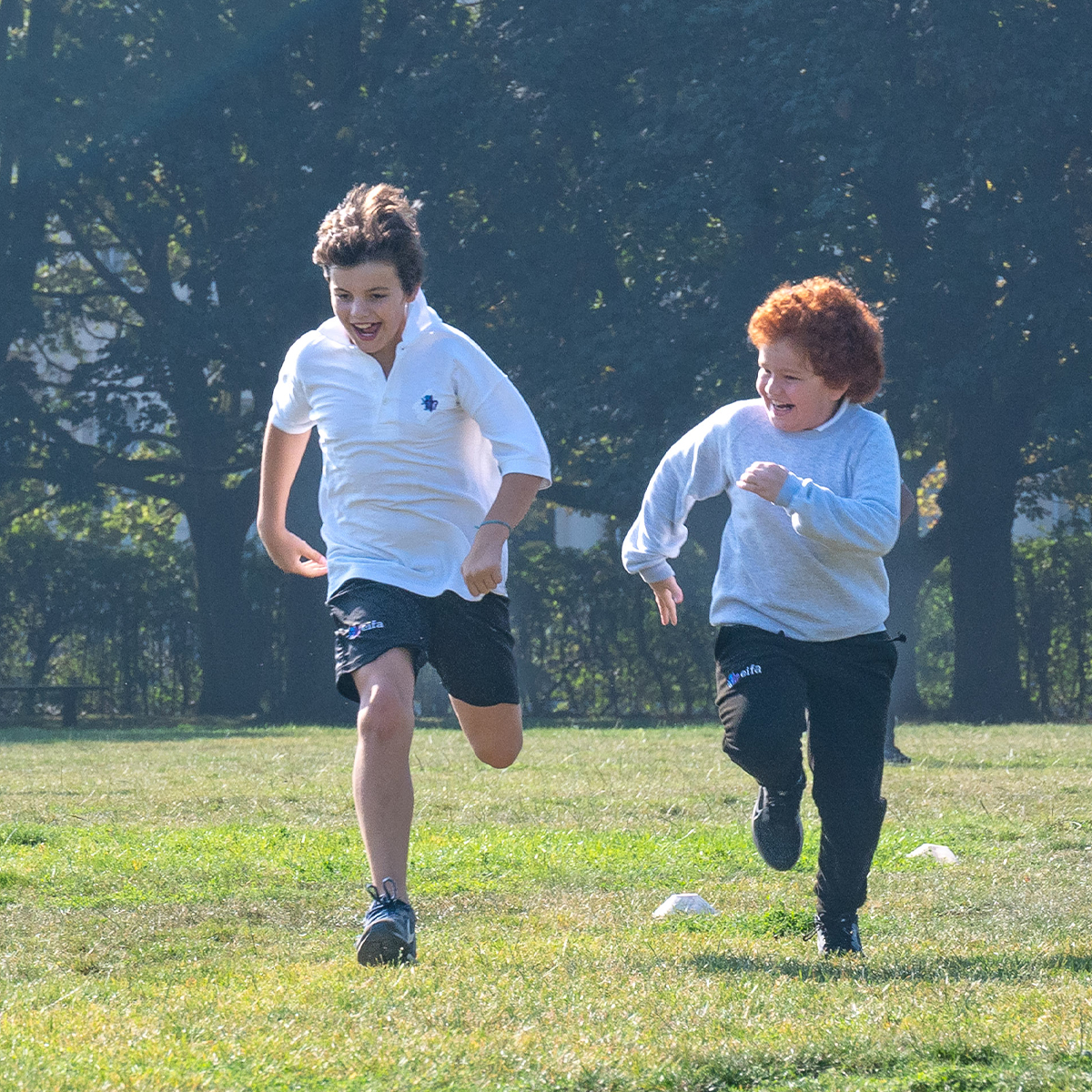 EIFA Two children, chatting in a mix of languages, are running across a grassy field, smiling and enjoying a sunny day. Both are wearing casual clothes as they play near a London school. Trees and a fence provide a charming backdrop to their carefree afternoon.