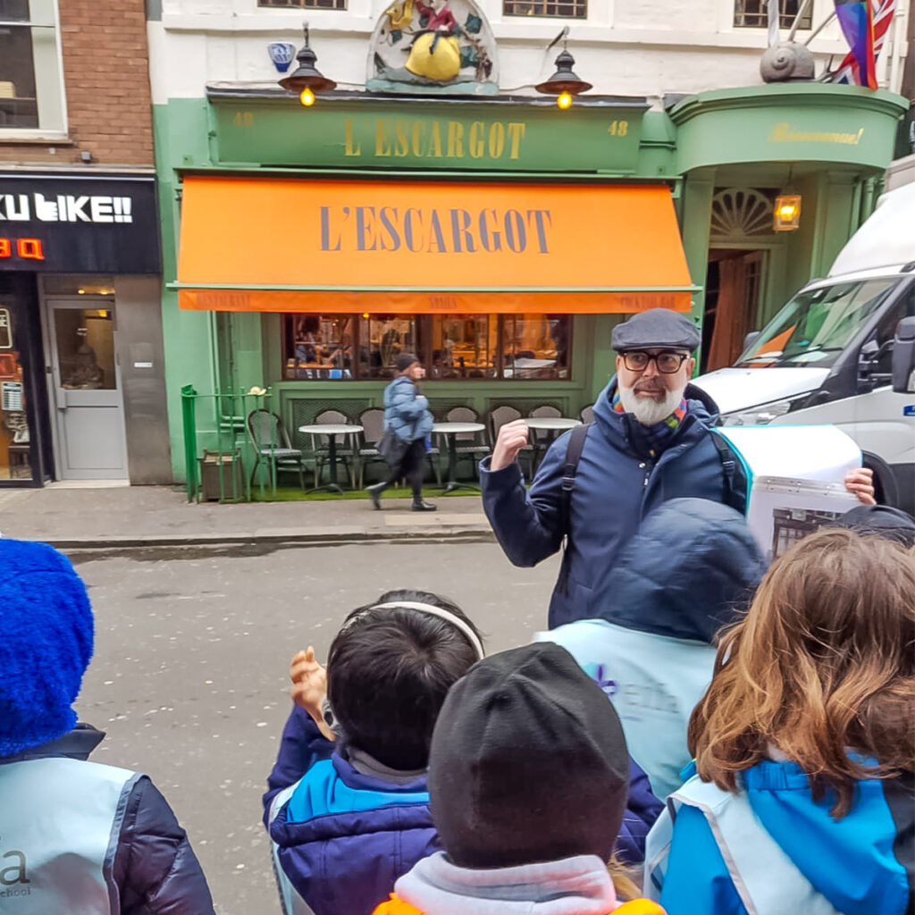 EIFA A group of children gathered around an enthusiastic guide holding a brochure outside the LEscargot restaurant. The restaurant features a bright orange awning and an ornate snail sculpture above it. The guide gestures energetically as the kids look on.