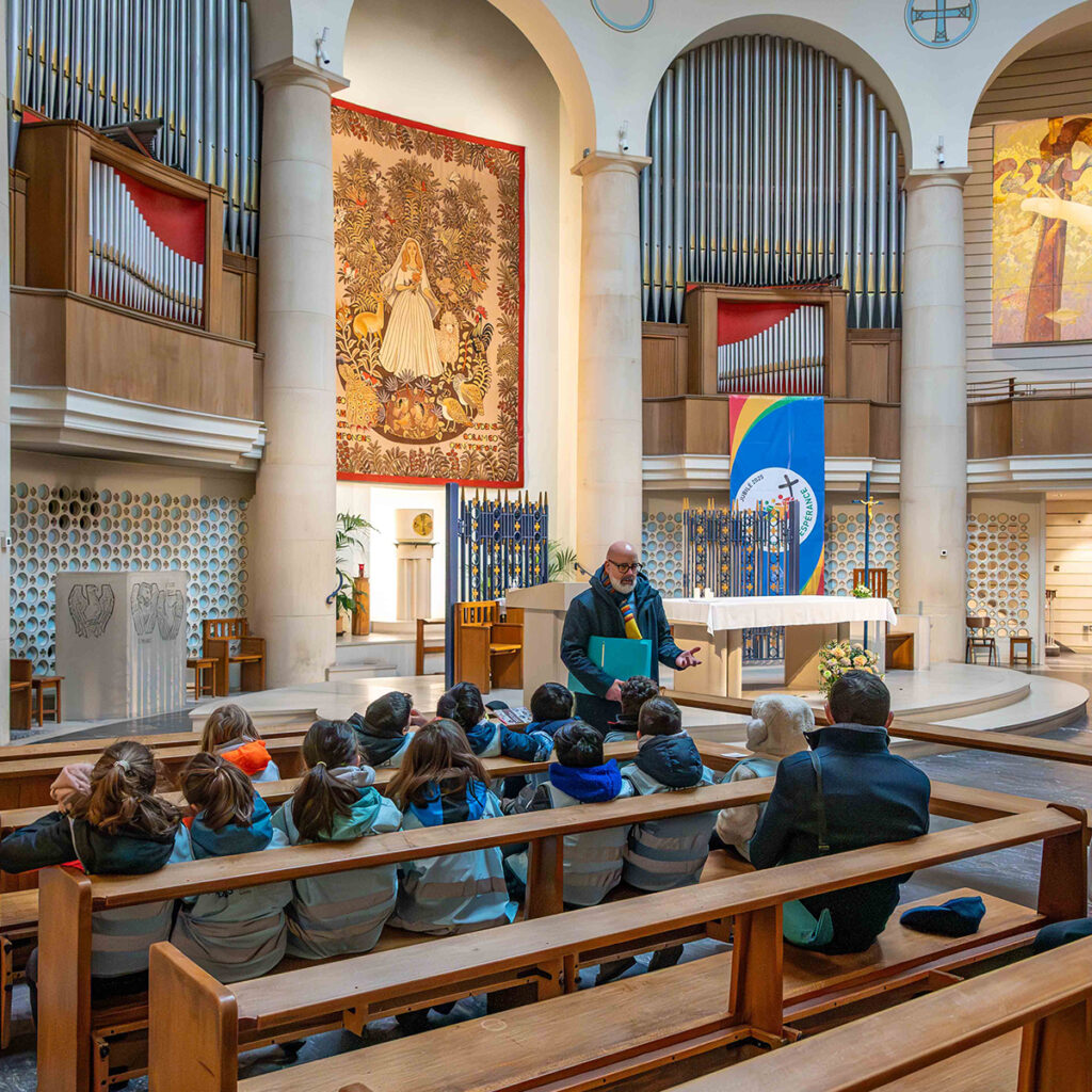 EIFA A group of children sit on wooden pews inside a church, listening to a man standing and speaking to them. The church interior features organ pipes, religious artwork on the walls, and a tapestry with a saintly figure.