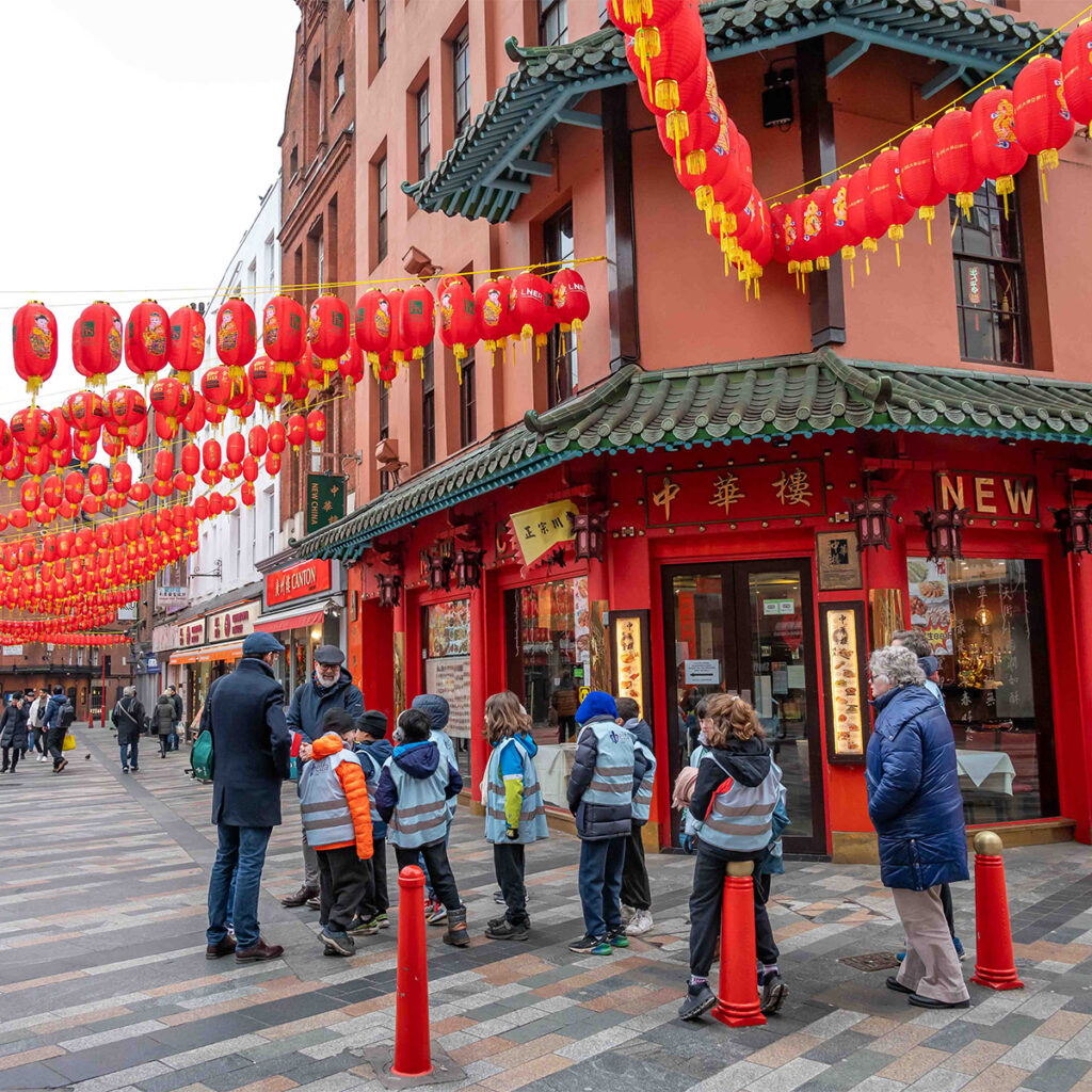 EIFA A group of people, including children, stand on a street in Chinatown. Red lanterns and festive decorations adorn the buildings. The architecture features traditional Chinese elements, and pedestrians are visible in the background.